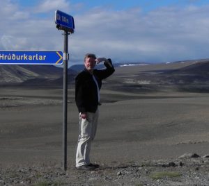 A man with short hair, shielding his eyes with his hand, by a signpost.