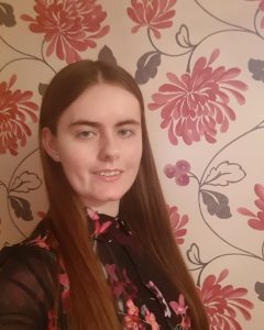 A yound woman with long, straight, dark hair, wearing a black shirt with pink flowers, standing in front of wallpaper featuring giant pink flowers.