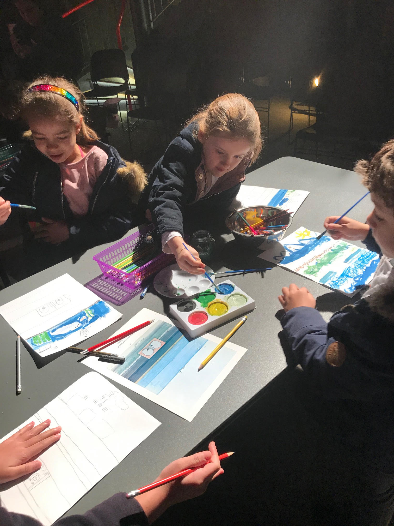 Several children at a table, painting. Dark background.