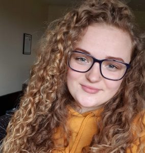 Head and shoulders of a smiling young woman with blonde curly hair and glasses.
