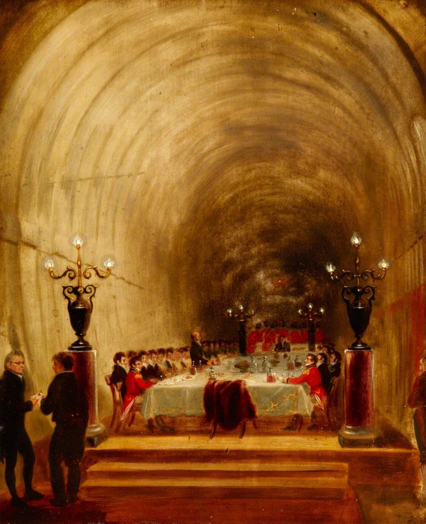 A banquet in a tunnel. There are two men front left, with a large table up some steps behind them. In the far background is a military brass band.