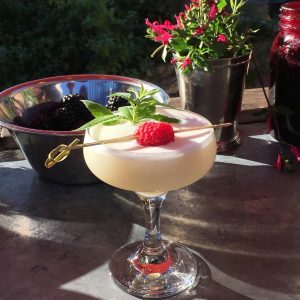 A creamy cocktail in a coupe glass decoated with a sprig of mint and a raspberry on a cocktail stick. There is a bowl of blackberries in the background and some pink flowers.