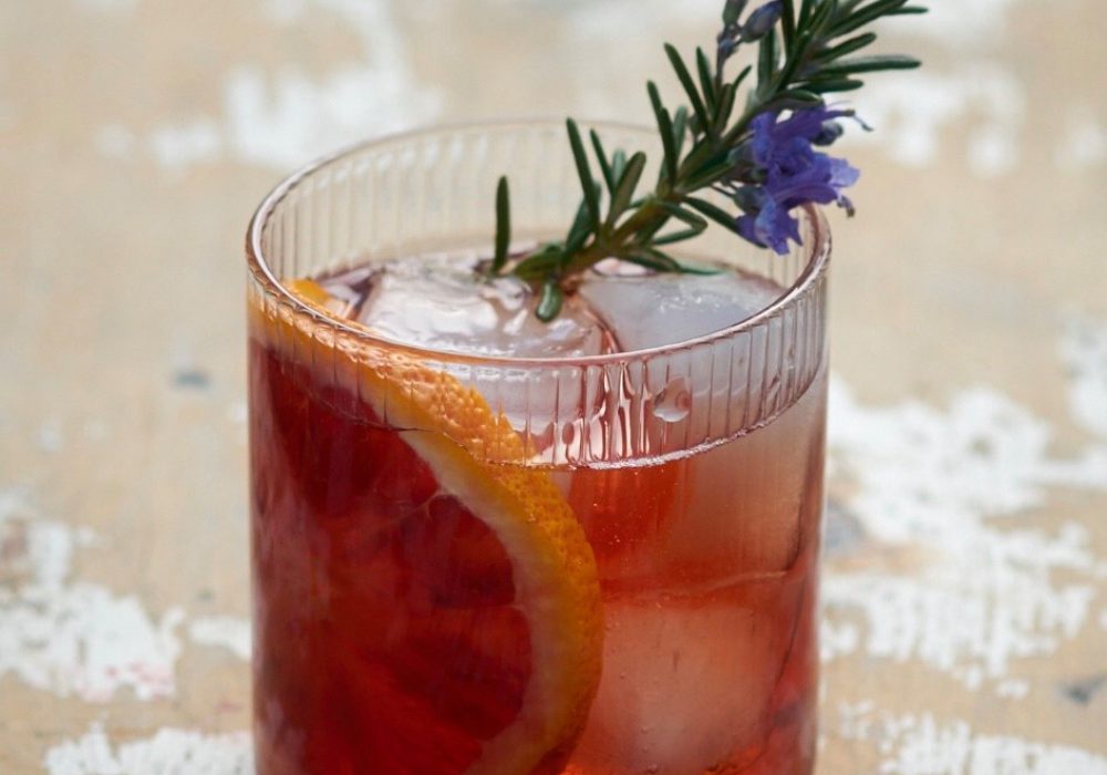 A cocktail in a short glass, decorated with a slice of orange and a flowering sprig of rosemary.