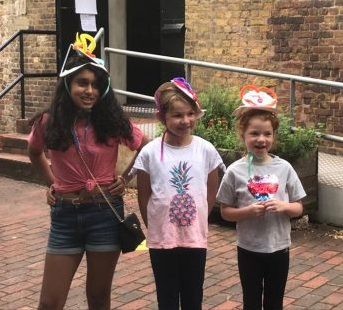 Three children wearing hats they have made from paper plates and ribbons and other bits and pieces, outside the Brunel Museum