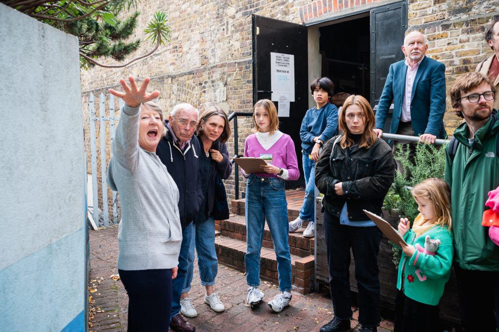 A female volunteer is talking to a group of people in front of the museum entrance. Two of the group, young girls, take notes on clipboards.