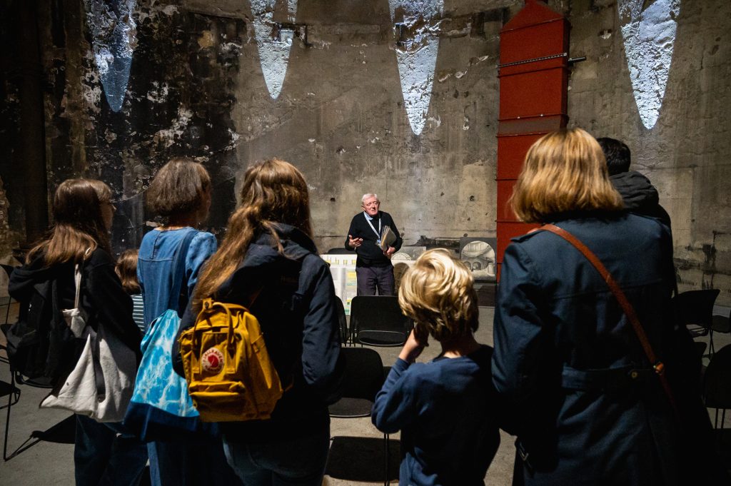 A male volunteer talks to a group of people at the base of the tunnel shaft. Behind him is the shaft wall. View from the back of the group.