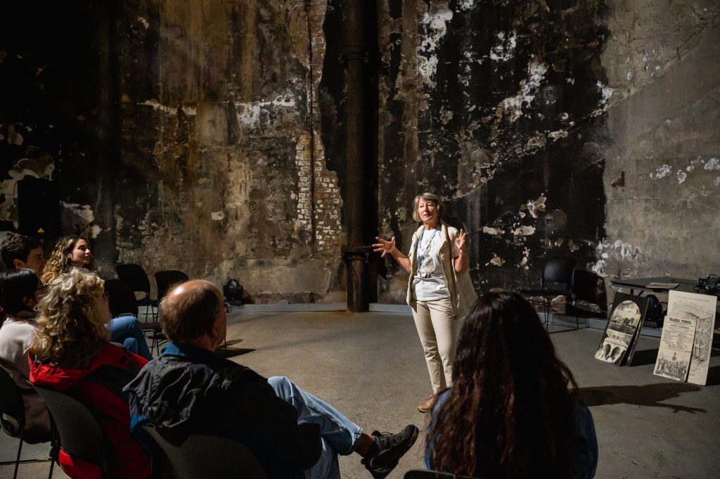 A female volunteer talks to a seated group of people at the base of the Tunnel Shaft. Behind her is the shaft wall.