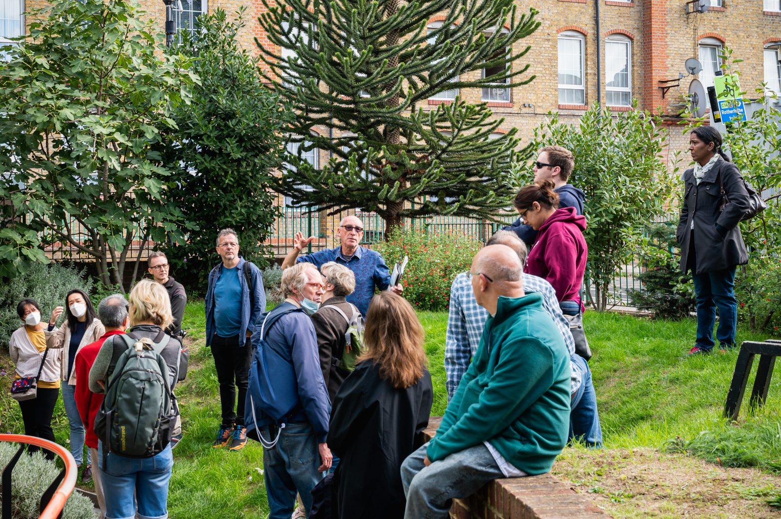 A volunteer, a man, talks to a group of people in the garden of the Brunel Museum. They are standing on a grassy bank. In the foreground, a man sits on a retaining wall.