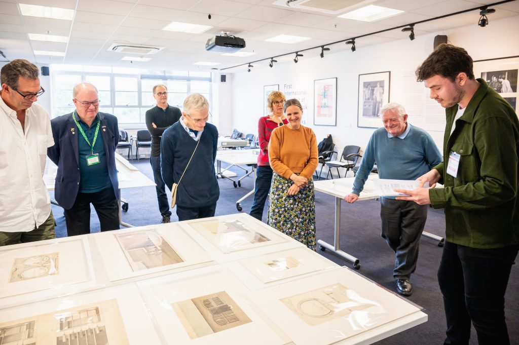 A group of people stand around a table. Laid out on the table are historical technical drawings. One of the group, a man, reads from a page of notes.