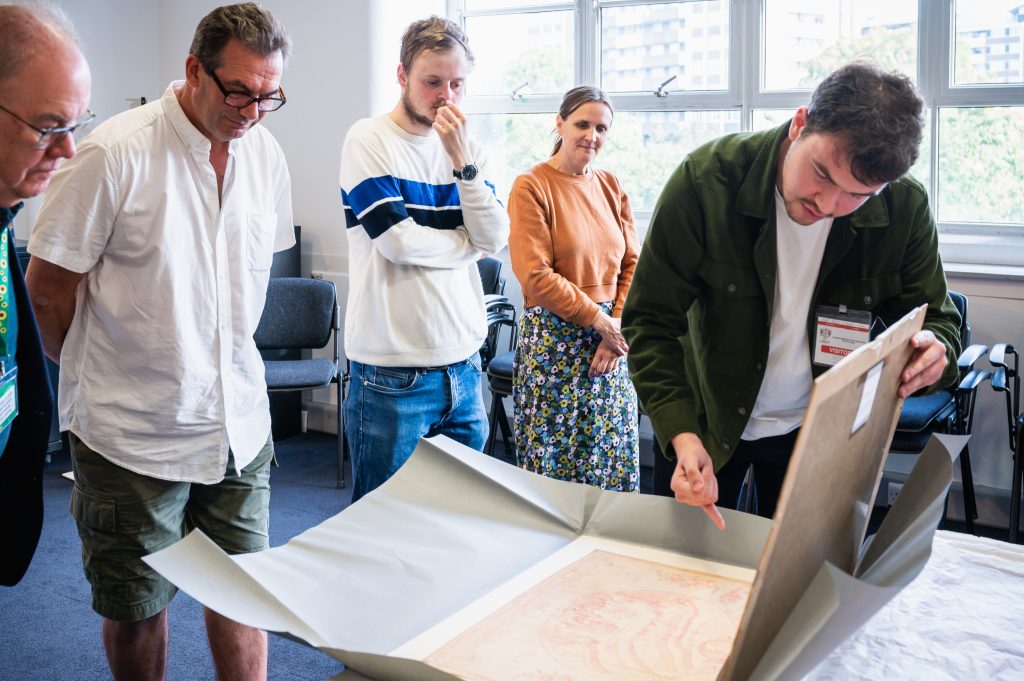 Five people look over a large, historic document. One of the group, a man, handles the document, gesturing to a part of it. He is wearing a lanyard labeled 'Visitor'.