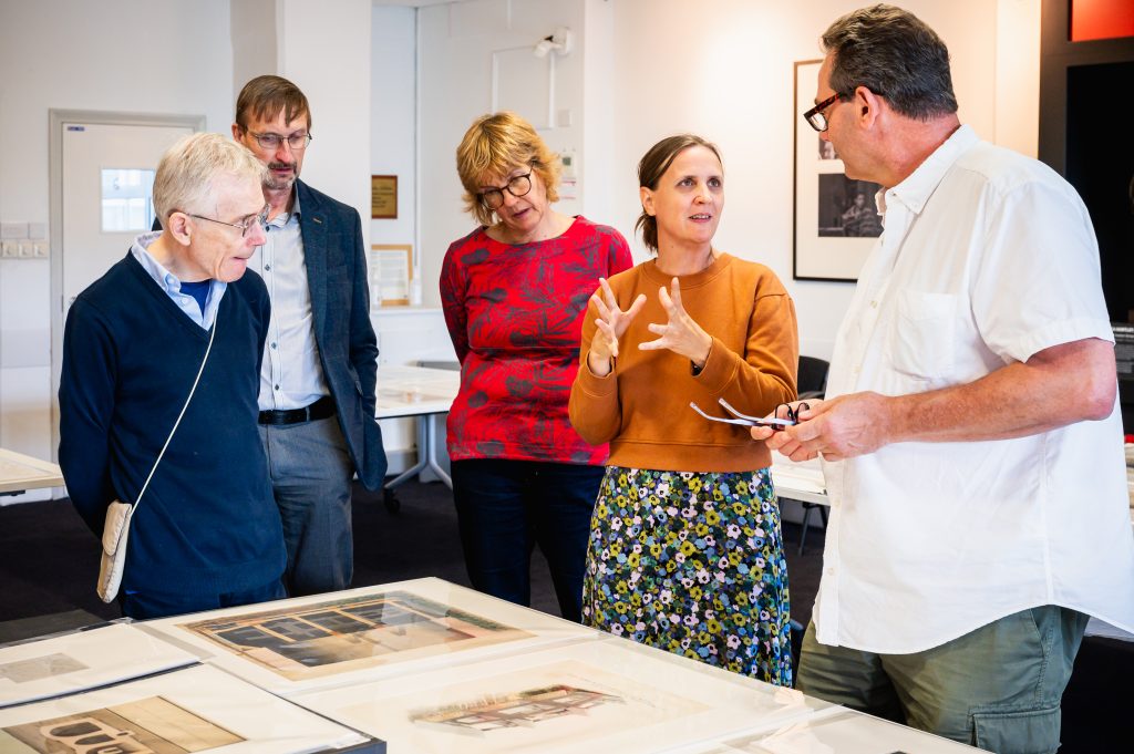 A group of five people look over historical documents which are laid out on a table. They are talking amongst themselves.