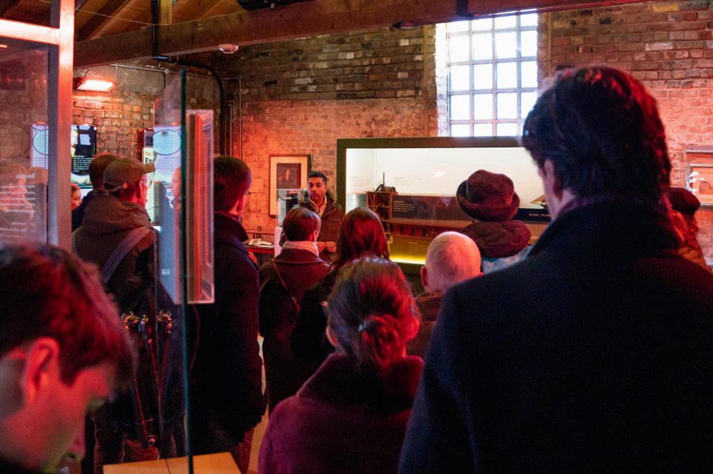 A volunteer talks to a group of people inside the Brunel Engine House. He is stood next to a display case holding a model of the tunnel shaft. View from the back of the group of people.