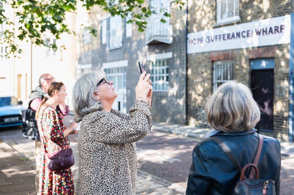 Four people looking at a row of buildings. A sign reading 'Hope (Sufferance) Wharf' hangs on the foremost building. One of the group, a woman, takes a picture of the buildings with her phone.