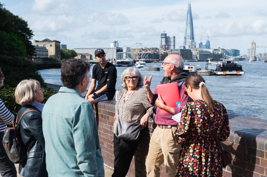 A volunteer gives a gives a talk to a group of people, while stood next to a wall on the river bank. In the background is the River Thames, the Shard and Tower Bridge.