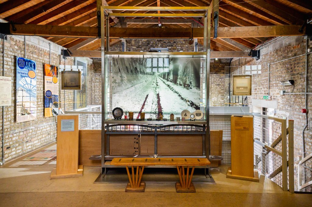 View of interior of the Brunel Engine House, looking from the back of the mezzanine towards display cabinet holding numerous small artefacts, in front of a photograph of the tunnel. Display panels are on the walls.