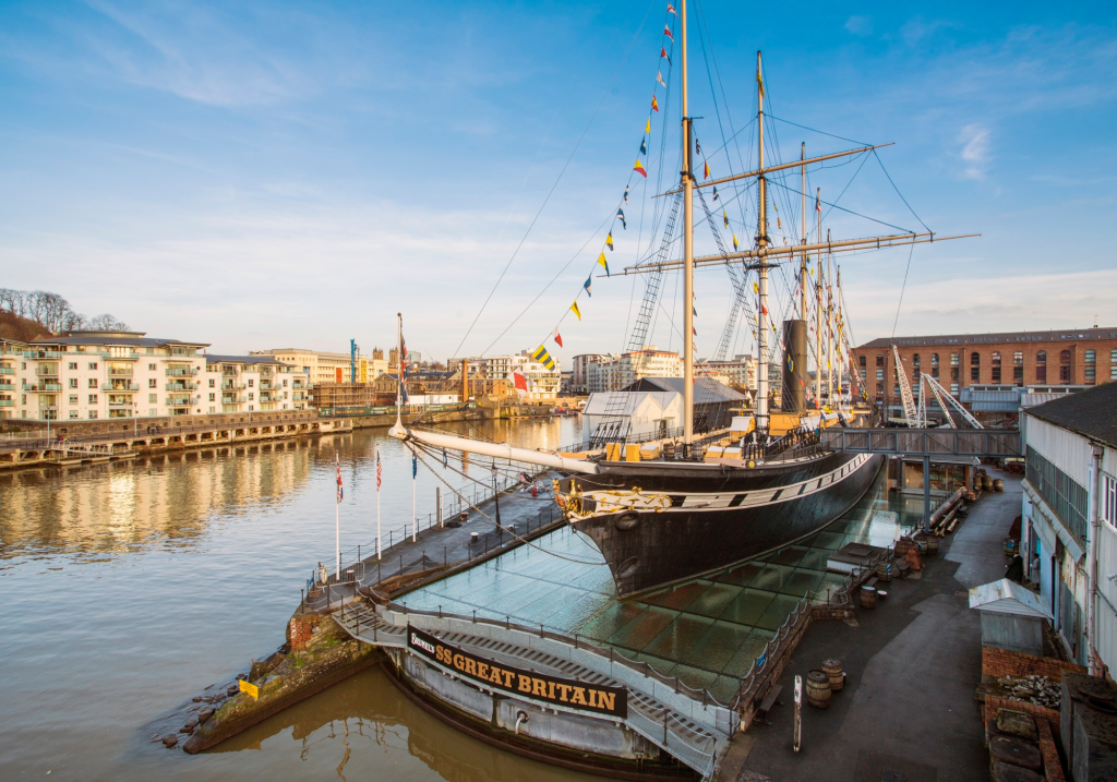 The ship SS Great Britain sits in a museum dry dock, surrounded by a glass floor around its hull. Coloured flags hang from the masts. A board reading 'Brunel's SS Great Britain' is on the front of the dry dock.