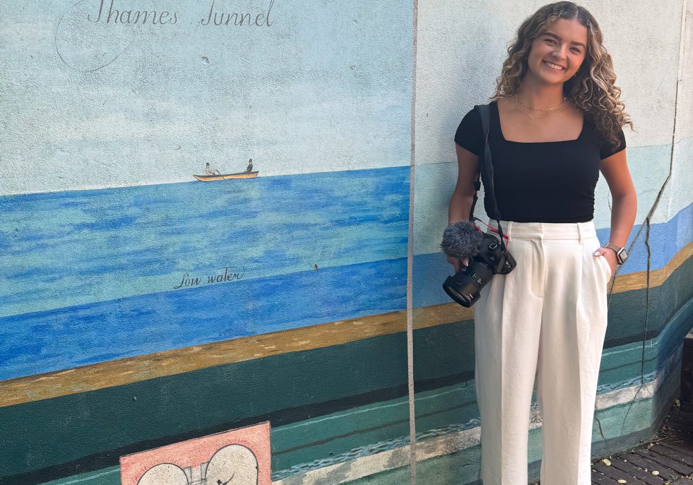 Image shows young woman holding a camera in front of a mural.