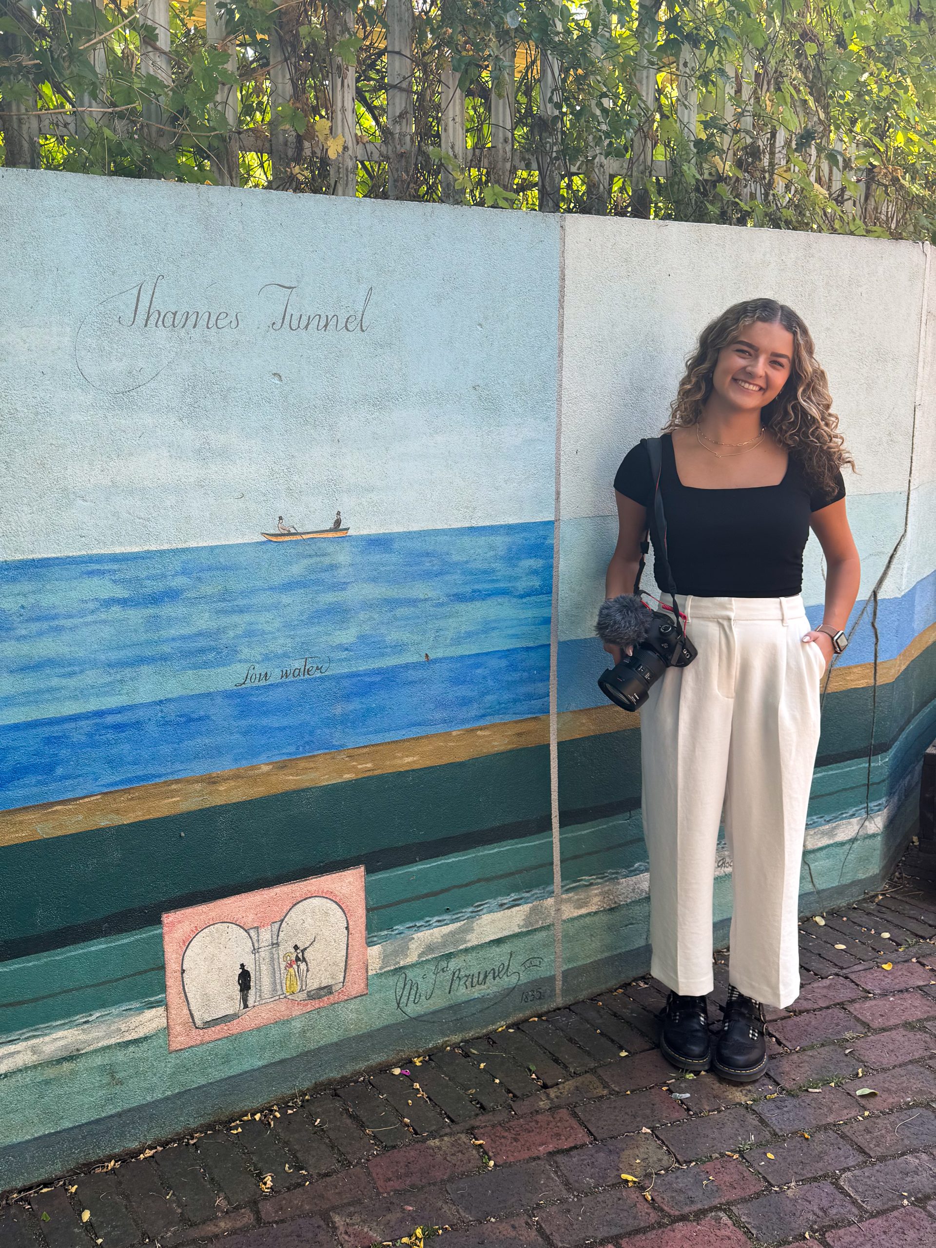 Image shows young woman holding a camera in front of a mural.