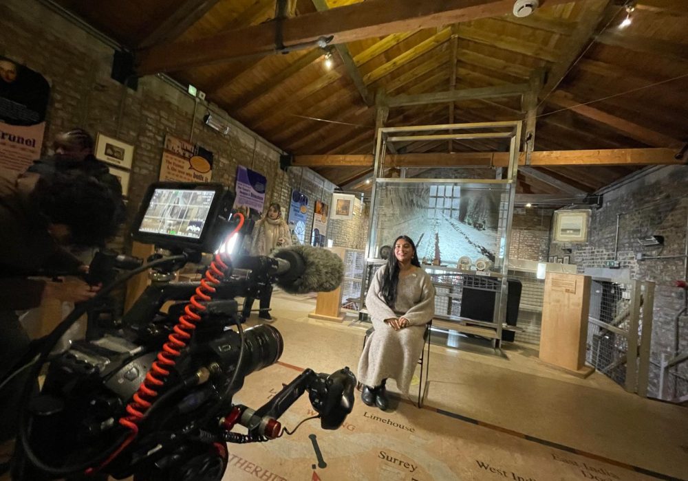 A South Asian woman in her 30s with very long black hair in a grey dress sits in a chair in front of a display case at the Brunel Museum. She is smiling and looking directly a large film camera with a boom microphone and attached display screen.