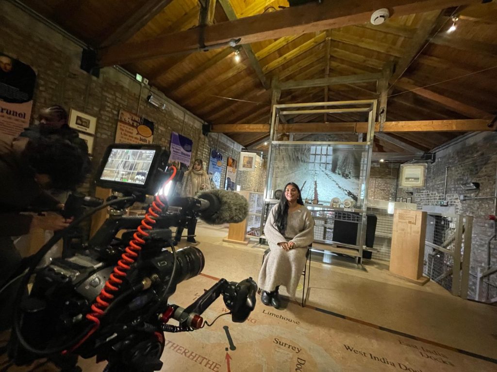 A South Asian woman in her 30s with very long black hair in a grey dress sits in a chair in front of a display case at the Brunel Museum. She is smiling and looking directly a large film camera with a boom microphone and attached display screen.