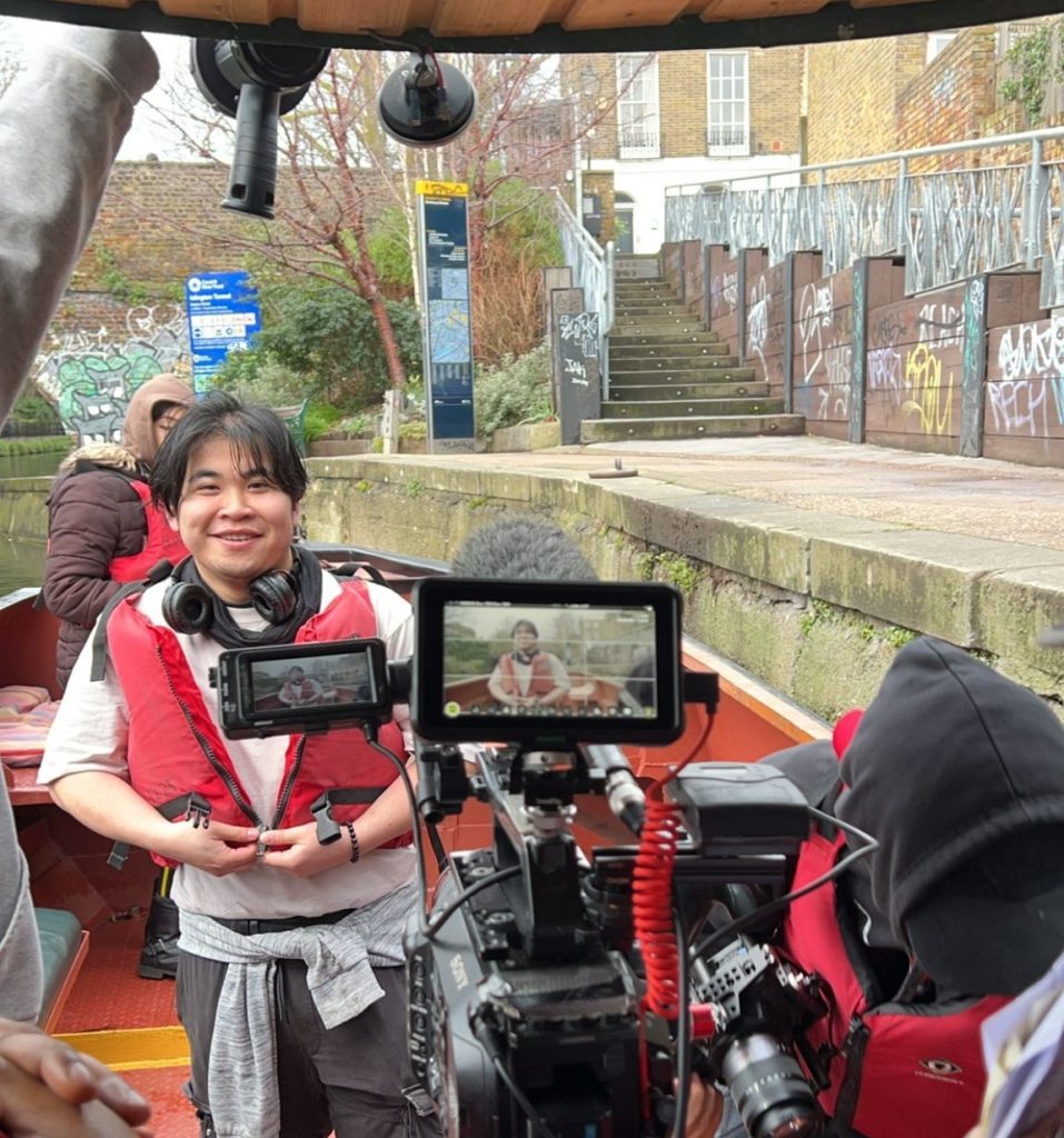 A young East Asian man in his early 20s stands on the front deck of a narrow boat on the London Canals. He is wearing a buoyancy aid and has over ear headphones around his neck. He is smiling and looking at a video camera. On top of the camera two screens capture and show an image of the man, repeating the image within the frame. 