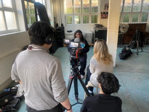 A young black woman in her early 20s sits facing the lens of video camera. Three young people stand and sit behind the camera. Only the backs of their heads are visible in the image.. 