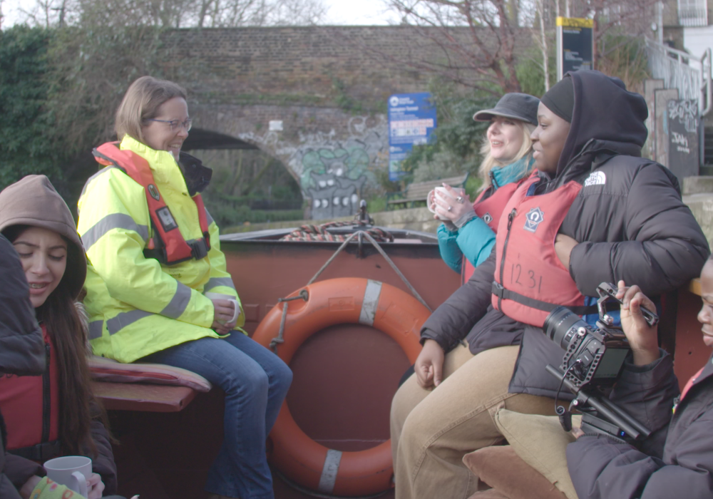 On the front of a foredeck of a narowboat, a White woman in her 40s sits opposite two young women in their early 20s. In front of them, three other young people talk while one of them holds a camera