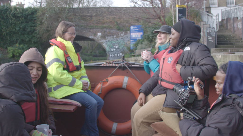 On the front of a foredeck of a narowboat, a White woman in her 40s sits opposite two young women in their early 20s. In front of them, three other young people talk while one of them holds a camera