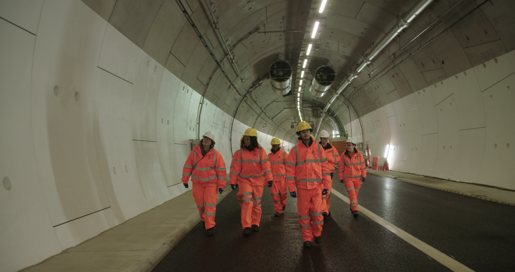 A group of six people of a mix of ages and genders walk through the Silvertown Tunnel while wearing orange personal protective equipment and white and yellow hard hats. 