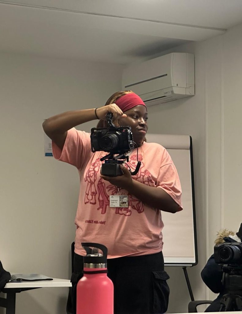 A young black woman wearing a pink t-shirt and a red scarf in her hair holds a video camera.