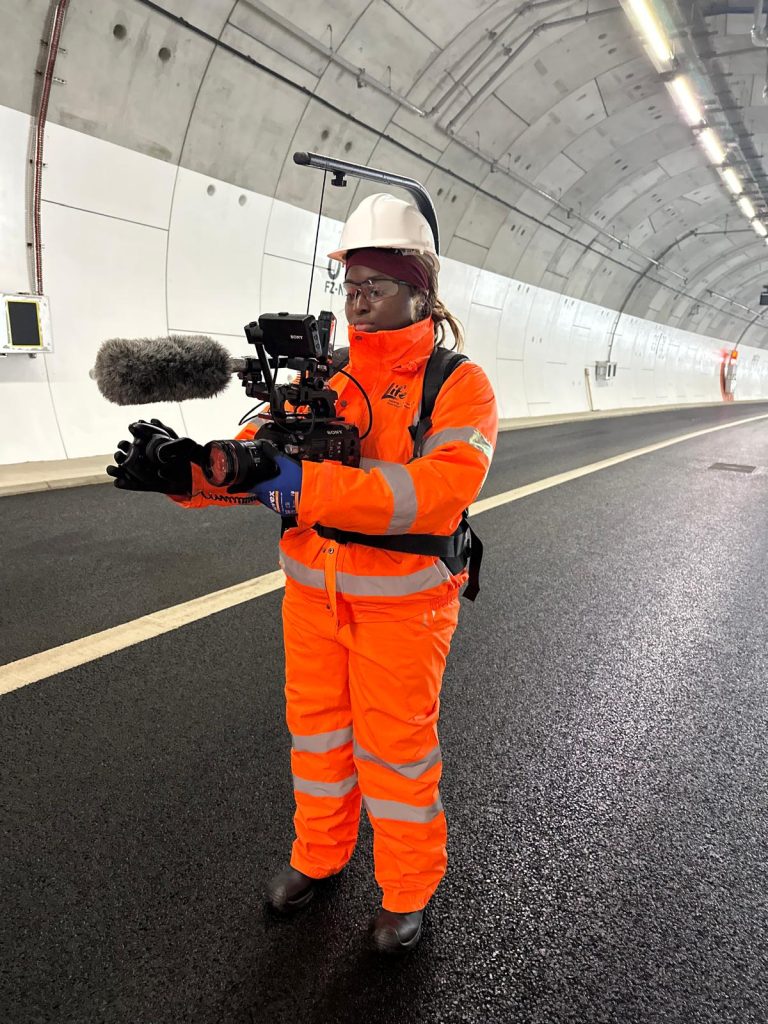 A young Black woman in her early 20s ear bright orange trousers and a jacket and a white hard hat. She wears a black rig supporting a large video camera that she holds in her hands. She's standing in a road tunnel. 