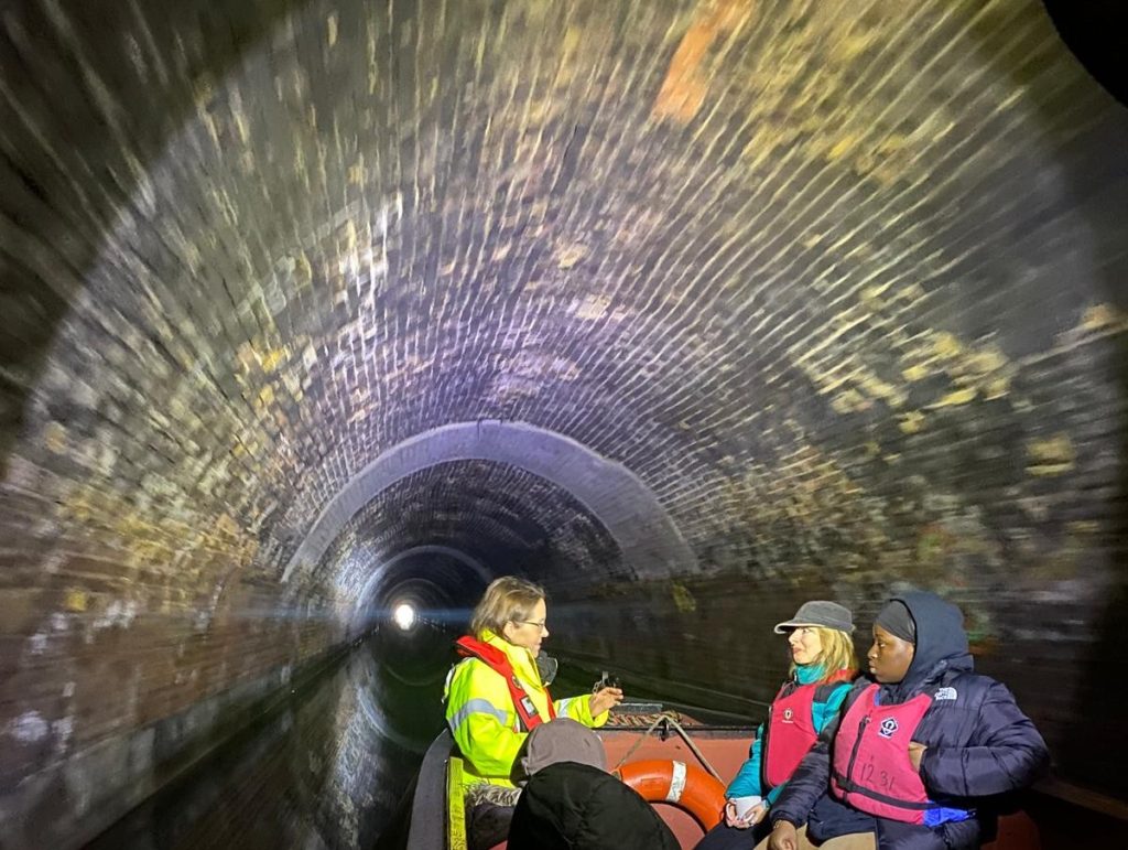 On the fore deck of a boat two young women in their early 20s sit opposite and talk to a woman in her 40s. They sitting on foredeck of a narrowboat, travelling through a canal tunnel with brown water in the bottom. A bright light illuminate the brickwork of the tunnel while in the distance, the opening of the end of the tunnel can be seen as a bright white circle. 