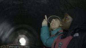 A young Black woman and a young white woman, both in their early 20s gaze up at the ceiling of a tunnel. They are surrounded by darkness except for a small circle of light from the end of the tunnel in the background